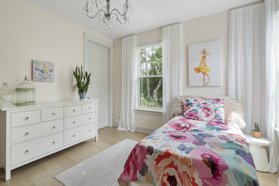 Modern bedroom with a colorful floral bedspread, white dresser, lush green plant, wall art, and large window with white curtains allowing natural light to brighten the space.
