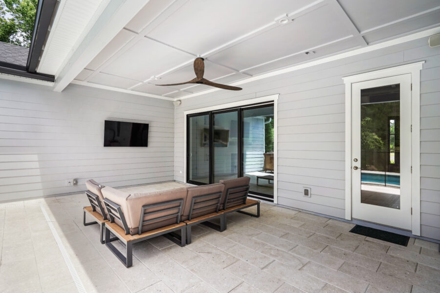 Modern covered porch with outdoor seating, ceiling fan, and sliding glass doors leading into the living space, featuring white siding walls and a view of a pool area.