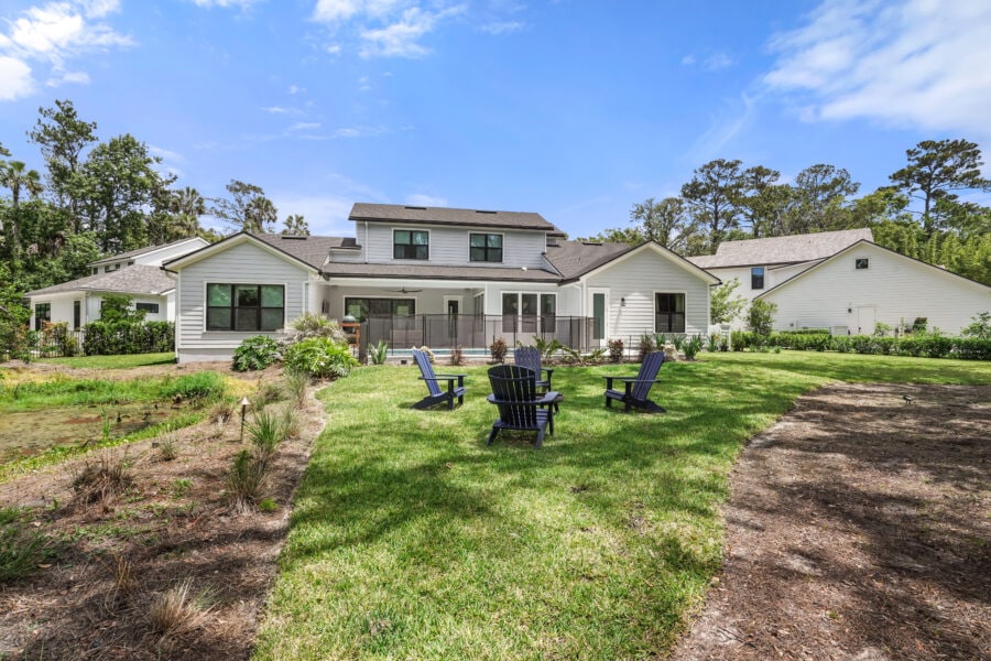 Spacious backyard with outdoor seating area, lush green lawn, garden beds, and a modern white house with large windows and a deck, surrounded by trees under a bright blue sky.