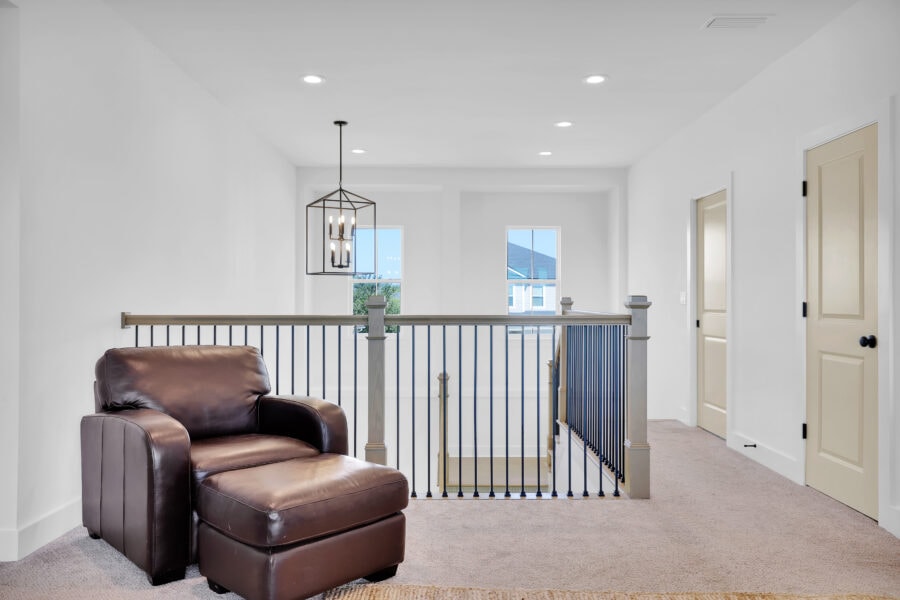 Modern bright upper hallway with beige carpet, white walls, black metal chandelier, and a leather armchair, overlooking a staircase with black metal balusters and windows providing natural light.