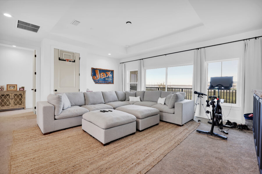 Bright living room with large windows, modern sectional sofa, exercise bike, and scenic views, showcasing comfortable and stylish home interior design by John Merrill Homes.