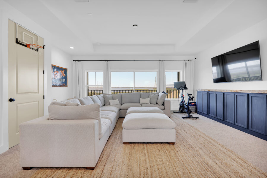 Spacious living room with large windows, white walls, beige sofa, and an entertainment area featuring a wall-mounted TV and exercise bike. Bright, airy, and perfect for family gatherings and relaxation.
