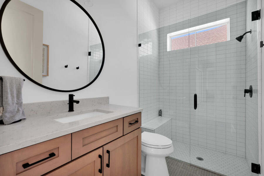 Modern bathroom with a white vanity, black fixtures, and a large round mirror, featuring a walk-in shower with white tiles, black hardware, and a high window for natural light.