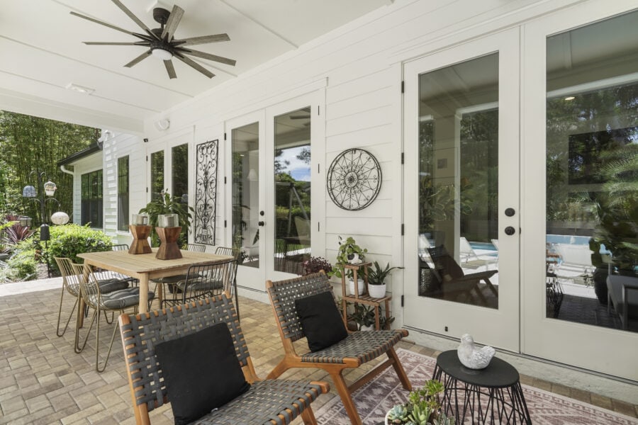 Bright outdoor patio with wooden dining table, wicker chairs with black cushions, potted plants, and decorative wall art, featuring a screened porch and lush garden background.