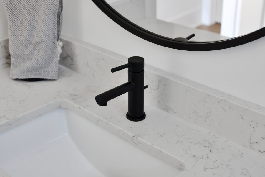 Sleek black faucet installed on a white marble bathroom sink with a large mirror and a patterned hand towel nearby. Modern and minimalist design typical of John Merrill Homes.