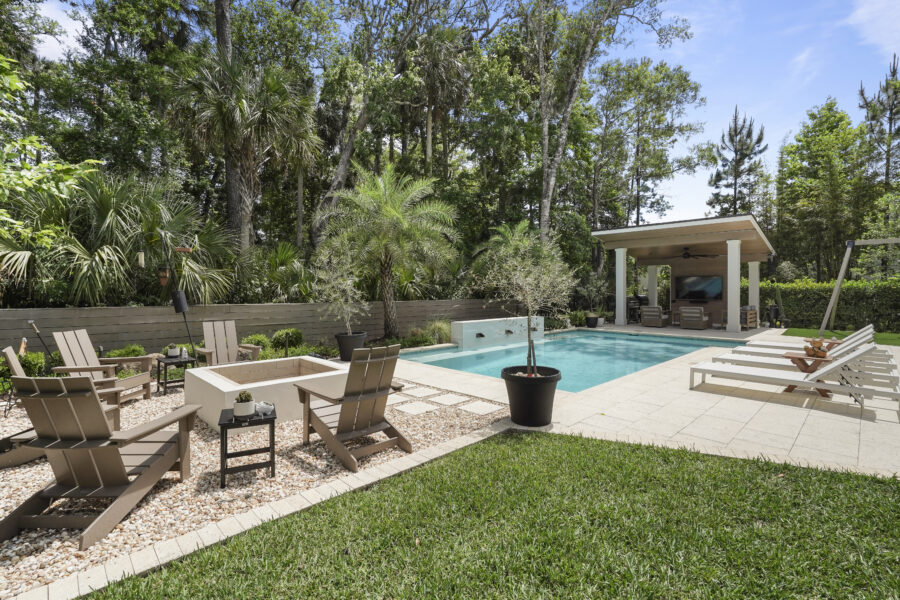 Luxurious backyard swimming pool area surrounded by lush greenery, comfortable lounge chairs, and a covered outdoor living space with a TV, creating an inviting space for relaxation and entertainment.