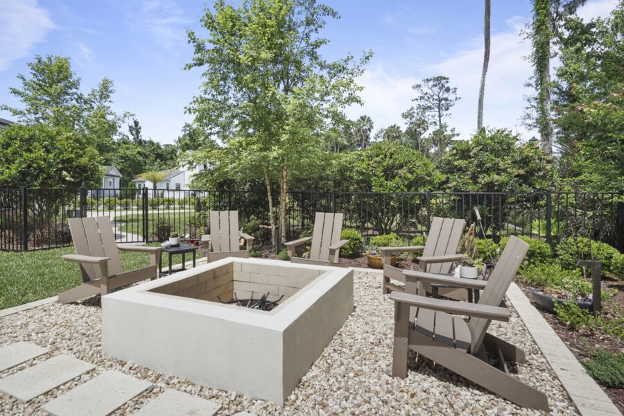 Cozy outdoor backyard fire pit area with Adirondack chairs, surrounded by lush greenery, trees, and a decorative black fence under a bright blue sky.