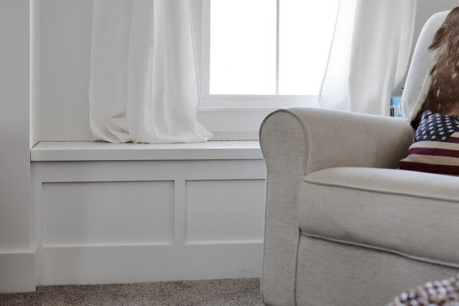 Comfortable beige armchair with patriotic throw pillow next to a large window with white curtains in a modern, bright living room.