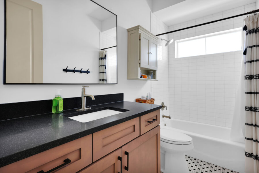 Bright modern bathroom featuring a black countertop vanity with a rectangular sink, wooden cabinetry, a wall-mounted mirror, white subway-tiled bathtub area with a horizontal window, and black-and-white patterned shower curtain.