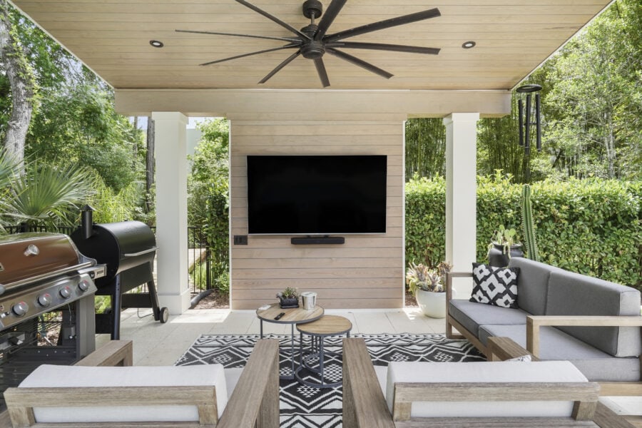 Modern outdoor living space with a mounted television, ceiling fan, comfortable seating, barbecue grill, and lush greenery surroundings for relaxing or entertaining.