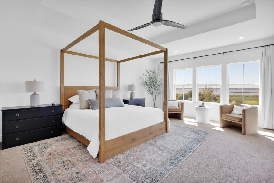 Bright and airy bedroom featuring a modern four-poster wooden bed frame, large windows offering scenic views, neutral-colored furniture, and elegant decor for a relaxing retreat.