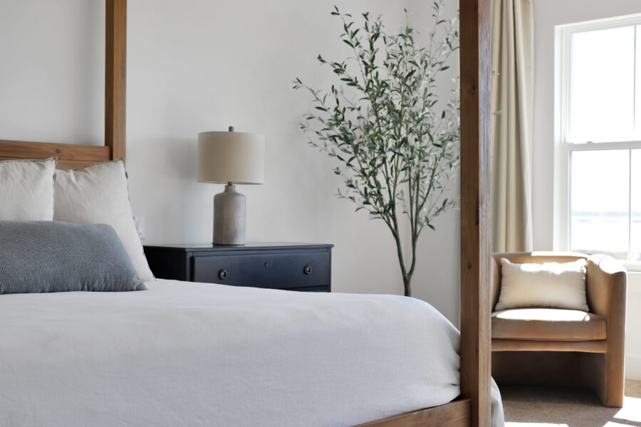 Bedroom with white bedding, a navy nightstand with a beige lamp, a large potted plant, cream curtains, and a beige armchair by the window at John Merrill Homes.