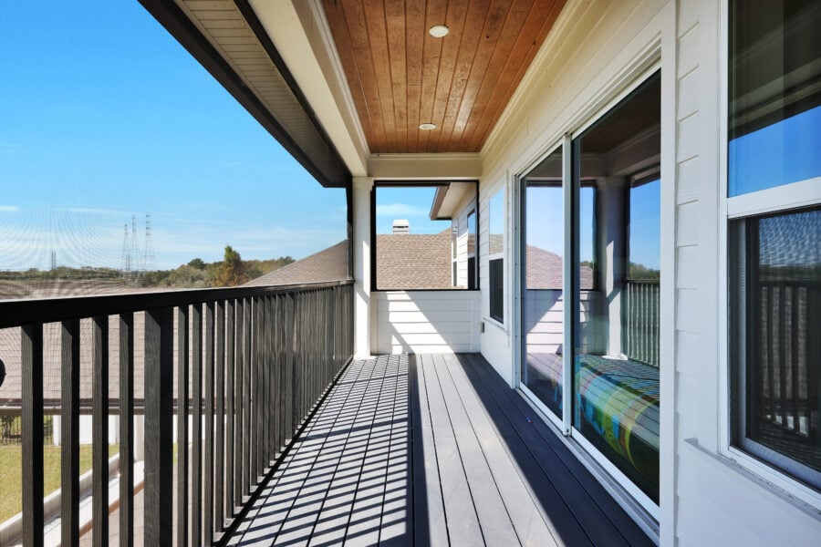A spacious balcony with a black railing, wooden ceiling, and sliding glass doors offering outdoor living space with a view of neighboring rooftops and trees under a clear blue sky.