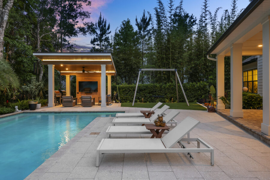 Relaxing backyard with a pool, outdoor lounge area, and a covered entertainment pavilion surrounded by lush trees at dusk.