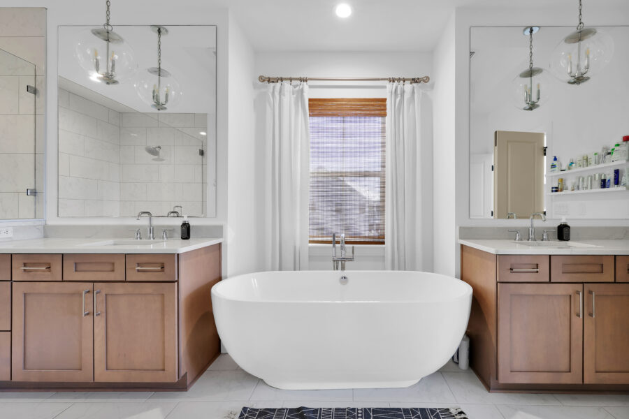 Luxurious modern bathroom with a central freestanding bathtub, double vanities with wooden cabinets, large mirrors, bright natural light, elegant fixtures, and a window with white curtains.