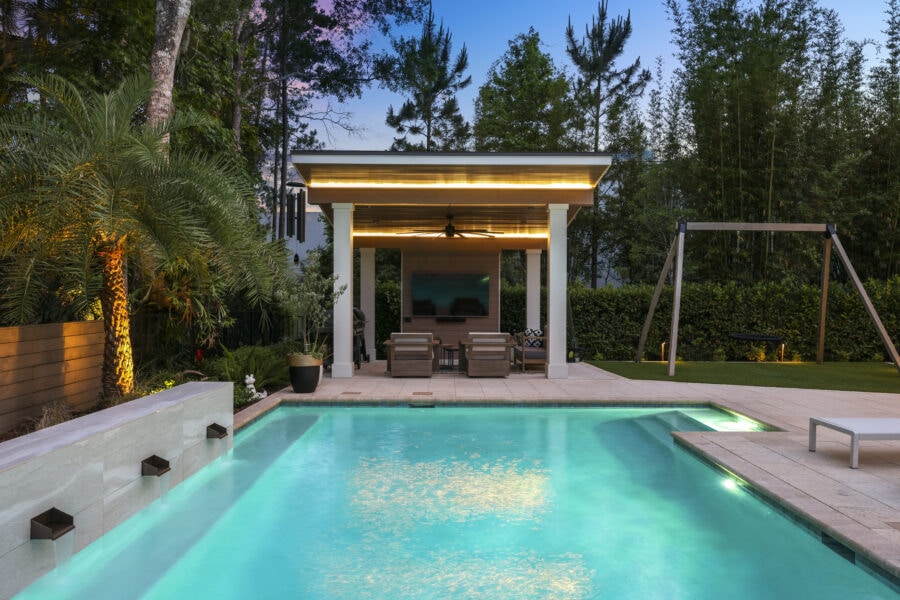 Luxurious backyard with a modern pool, outdoor entertainment area with TV, seating, lush greenery, and children's swing set, showcasing stylish outdoor living designed by John Merrill Homes.