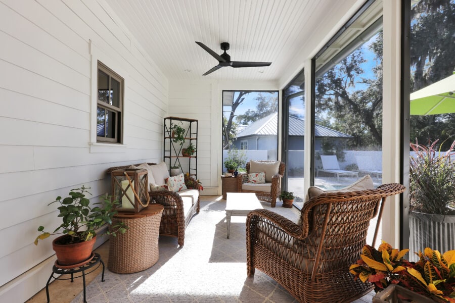 Bright sunlit screened porch with wicker furniture, potted plants, and cozy cushions, offering a relaxing outdoor space with views of trees and neighboring houses.