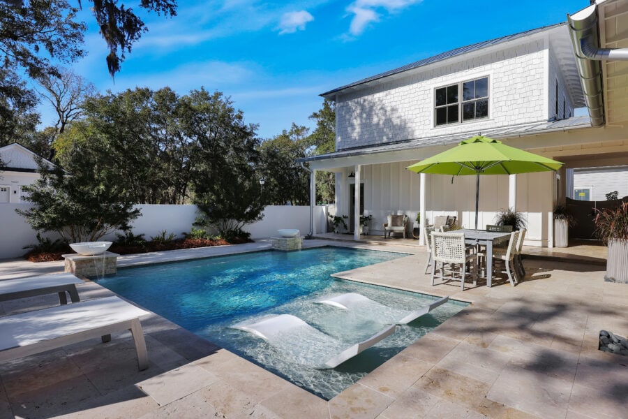 Luxury backyard swimming pool with spa jets, outdoor dining area with green umbrella, and modern patio furniture in a private home yard under a bright blue sky.
