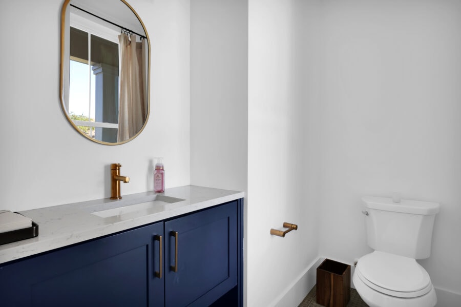 Modern bathroom featuring a white marble countertop vanity with navy blue cabinets, a gold faucet, a large mirror, a toilet, and natural light coming through a nearby window.