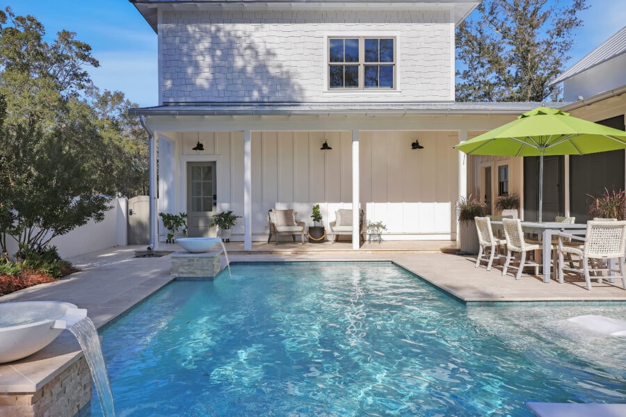 Luxurious backyard featuring a modern in-ground swimming pool, outdoor seating area with cushioned chairs, a green patio umbrella, and stylish decor against a white house exterior with large windows.