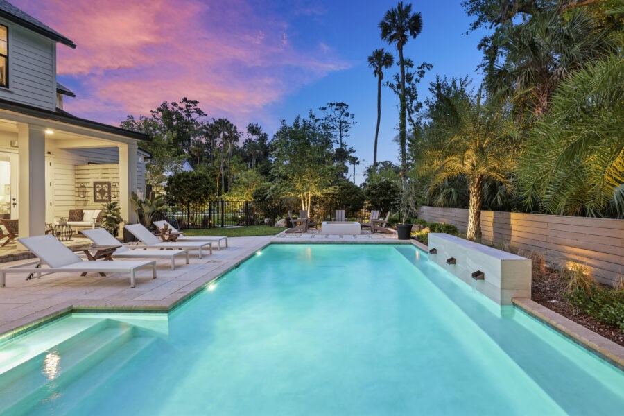Luxurious outdoor swimming pool at twilight with lounge chairs, lush tropical landscaping, cozy seating area, and vibrant sunset sky, showcasing elegant backyard design by John Merrill Homes.