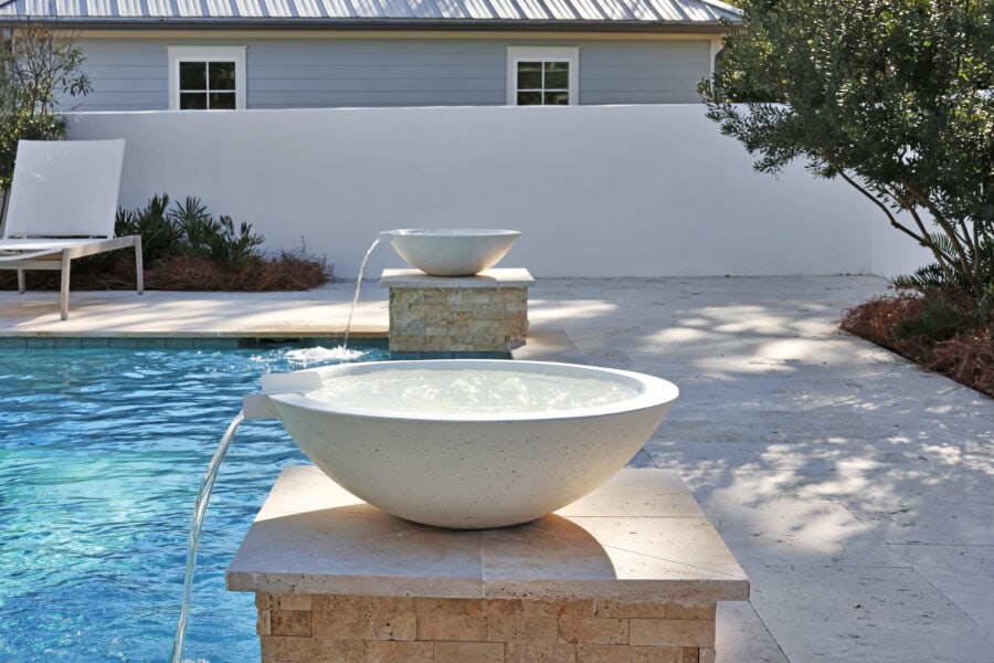 A modern backyard pool area featuring a stone fountain with two ceramic bowls, lush greenery, and a white privacy wall, providing a serene outdoor space perfect for relaxing and entertaining.