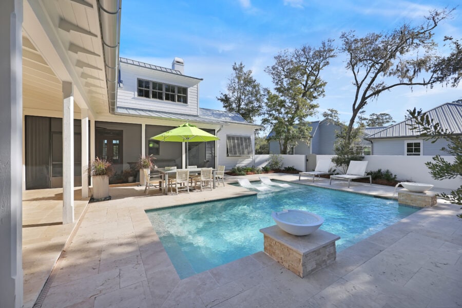 Luxurious outdoor swimming pool with lounge chairs, a dining area with a table and umbrella, surrounded by a modern patio, trees, and a stylish house with large windows and white siding.