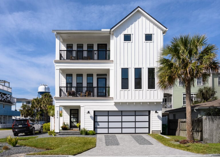 Modern white coastal-style home with vertical paneling, multiple balconies with black railings, large black-framed windows, and a landscaped front yard with a paved driveway.