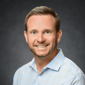 John Merrill Homes sales professional, smiling with a professional headshot in a light blue checkered shirt against a dark gray background.