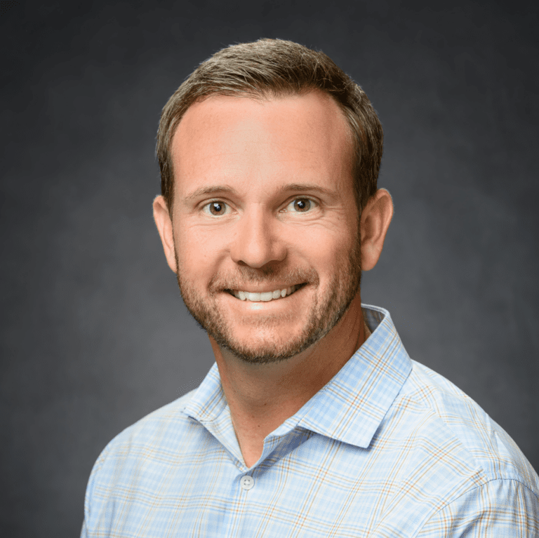 John Merrill Homes sales professional, smiling with a professional headshot in a light blue checkered shirt against a dark gray background.