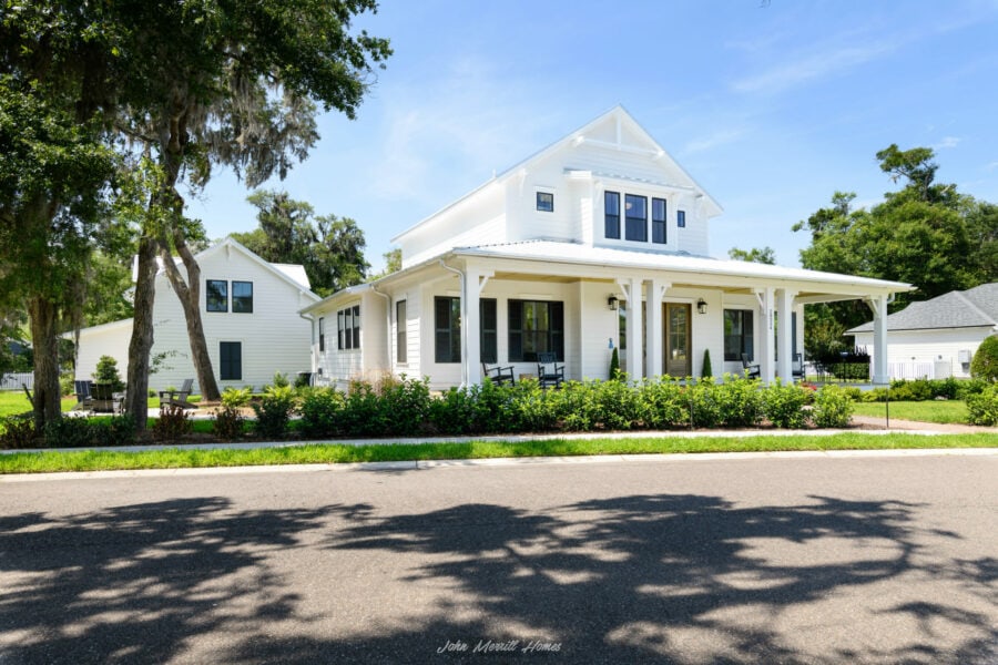 White modern farmhouse-style home with a large front porch, surrounded by lush green landscaping and tall trees on a sunny day.