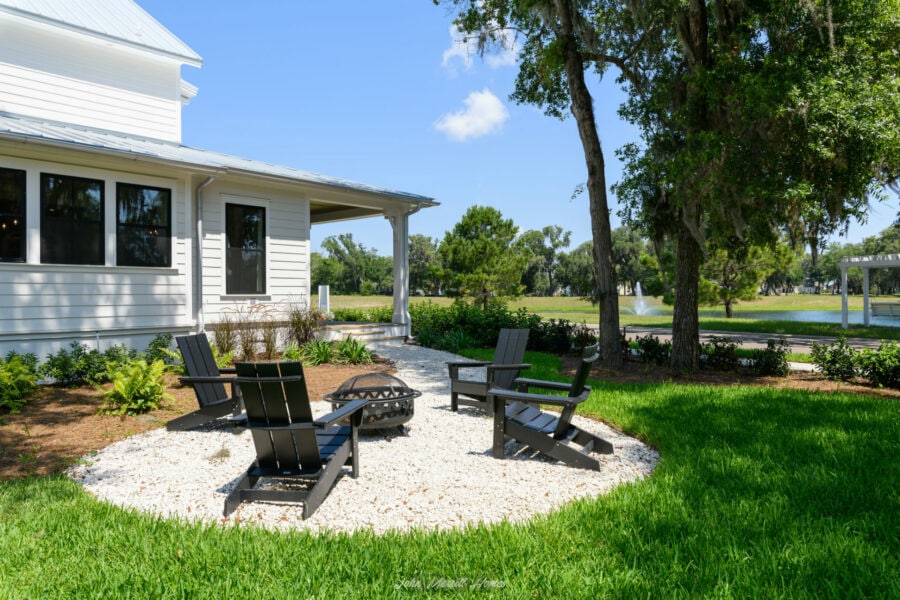 Fire pit area with black Adirondack chairs on white gravel, surrounded by lush green lawn and trees, overlooking a pond with a fountain at John Merrill Homes.