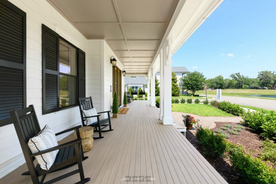 Porch with black rocking chairs, decorative pillows, and potted plants outside a white house with black shutters, overlooking a lush green lawn and street on a sunny day.