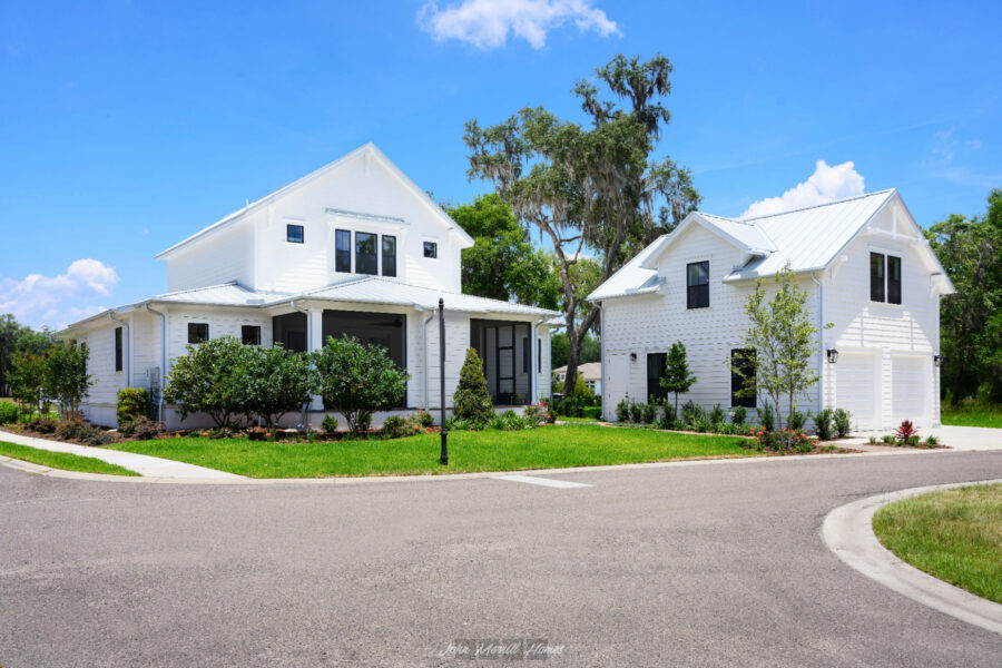 Modern white farmhouse-style homes with black accents, surrounded by lush landscaping and mature trees, showcase new construction homes by John Merrill Homes in a beautiful neighborhood.