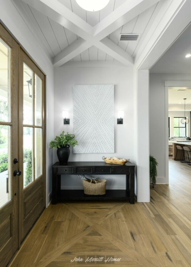Bright entrance hall with natural wood double doors, white walls, a black console table with a potted plant and wicker basket, and a vaulted ceiling with white wooden beams.