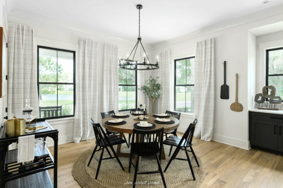 Round wooden dining table with black chairs in a bright, modern dining room with large windows, white walls, and black accents.