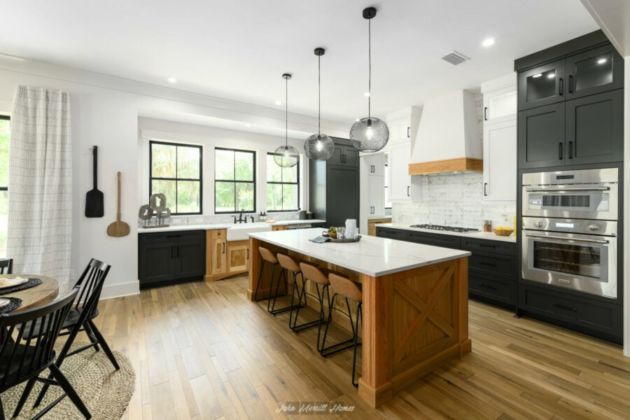 Spacious modern kitchen with black and white cabinetry, a large central island with wooden accents, pendant lighting, and large windows providing natural light.
