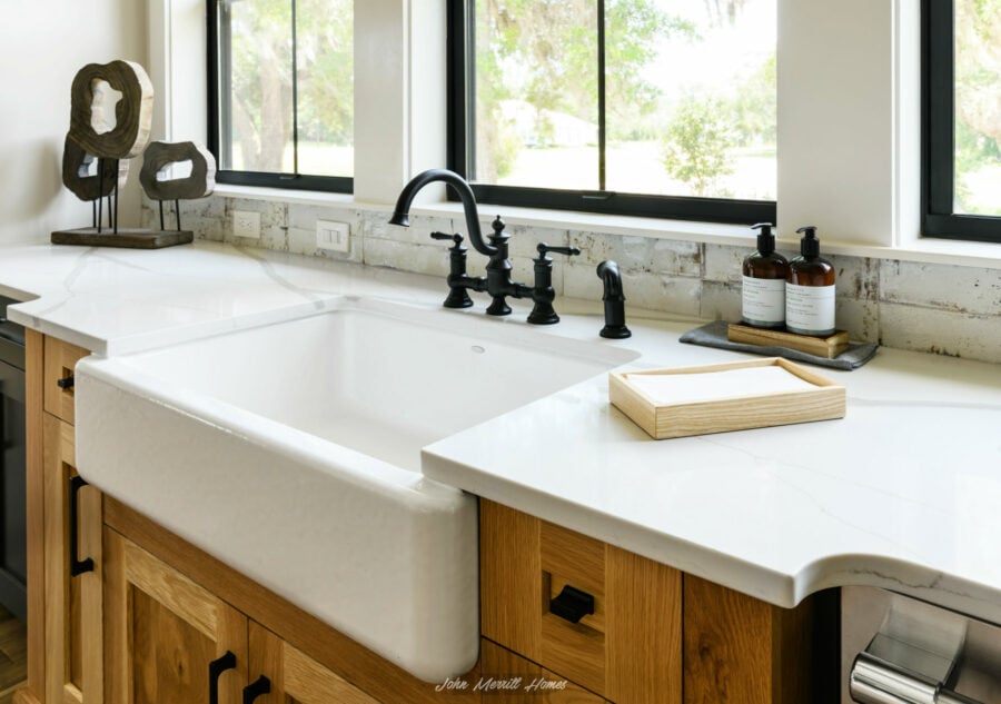 Rustic farmhouse kitchen sink with black faucet and soap dispensers, natural wood cabinetry, and large windows bringing in natural light.