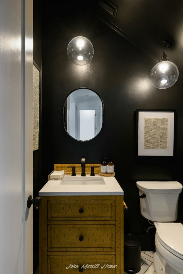 Modern bathroom with a black wall, a white vanity with a yellow textured finish, black fixtures, a round mirror, and stylish pendant lights.