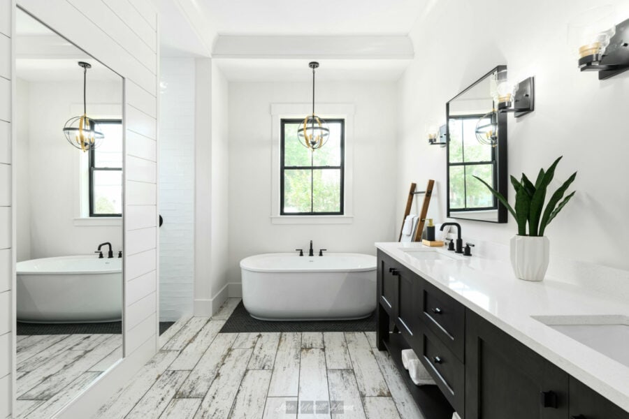 Bright modern bathroom with a freestanding bathtub, black fixtures, and a double vanity with black cabinets, white countertops, and decorative lighting, featuring natural light from windows.