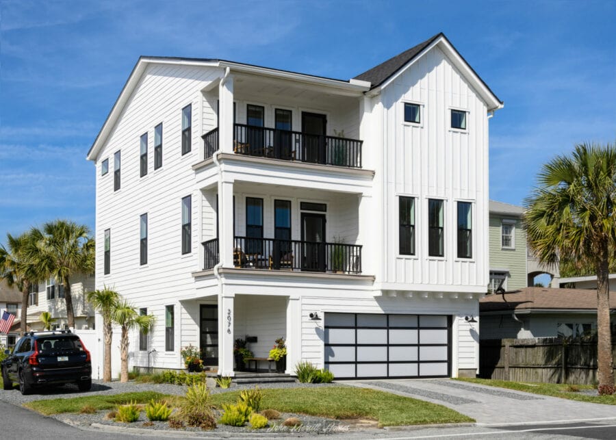 Modern white multi-story home with black window frames and railing balconies, surrounded by palm trees, under a bright blue sky.