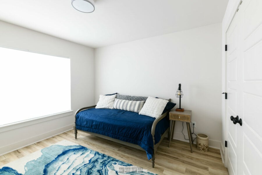 Modern bedroom with white walls, a large window letting in natural light, and a cozy bed with a blue quilt, decorative pillows, and a small side table with a lamp.