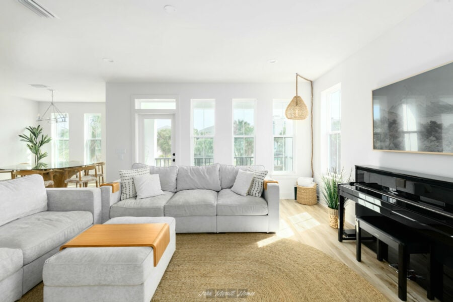 Bright and airy living room with large windows, modern gray sofas, a black piano, and natural decor elements, offering a welcoming and stylish home interior.