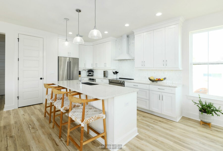 Bright white modern kitchen with cabinetry, stainless steel appliances, and an island with wooden bar stools, featuring natural light from a large window and a light wood floor.