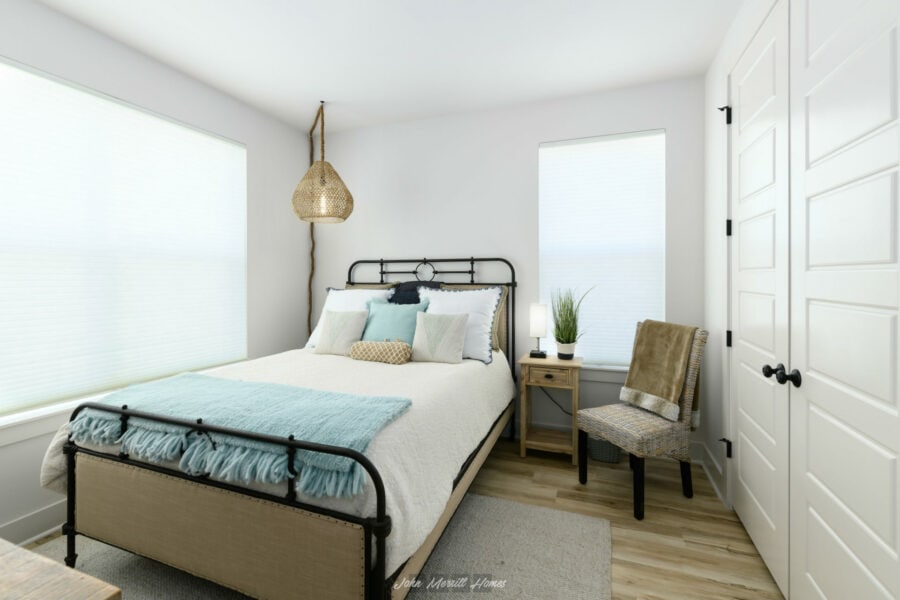 Bright bedroom with white walls, large windows with blinds, a black metal bed frame, cozy bedding, decorative pillows, a wooden nightstand with a lamp and plant, and a woven chair, creating a modern, inviting, and airy space.