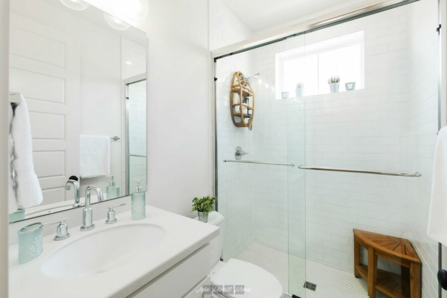 Bright modern bathroom with white tiles, a glass shower enclosure, and minimalist decor, featuring natural light from a window and a small wooden bench for seating or storage.