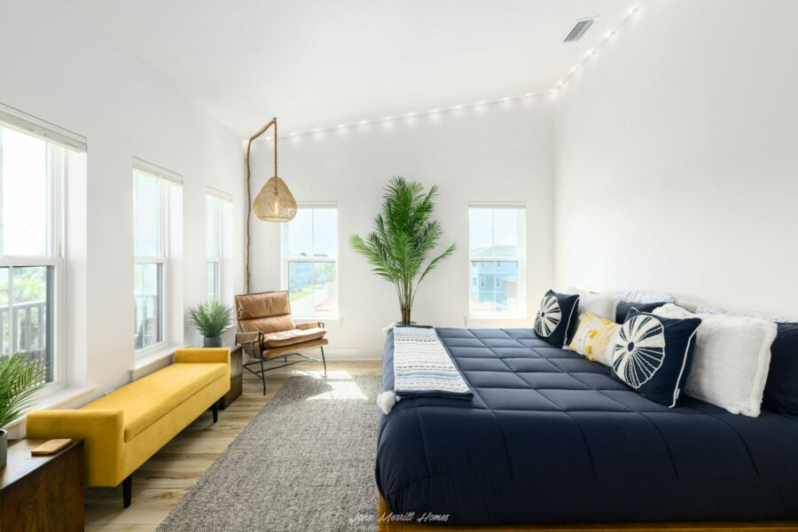Bright bedroom with large windows, a navy bed with decorative pillows, a yellow bench, a leather armchair, a tall potted plant, and soft natural lighting creating a cozy, modern space.