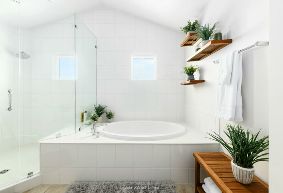 Bright modern bathroom with white tiled walls, a large corner bathtub, glass-enclosed shower, and decorative green plants on wooden shelves and a bench. Natural light enters through small windows, creating a fresh and relaxing atmosphere.