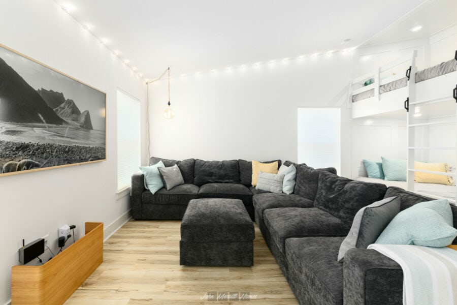 Cozy living room with a large dark gray sectional sofa, colorful throw pillows, a wall-mounted black-and-white landscape photo, and a built-in bunk bed with a ladder for children. The space features white walls, natural light, and warm wood flooring.