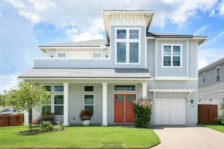 Bright, modern two-story house with light blue and white exterior, featuring large windows, a red front door, lush green lawn, small trees, and a driveway under a partly cloudy sky.